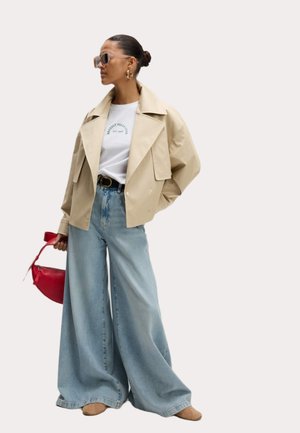 Beige cropped jack, witte T-shirt met groene print, lichte blauwe wijde broek, en bruine schoenen; houdt een rode handtas vast. Vierkante zonnebril.