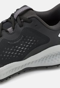 Chaussure de sport noire avec une tige en mesh texturé, un bout renforcé et une semelle blanche rembourrée. Comprend un lacet réfléchissant et des accents sur les côtés.