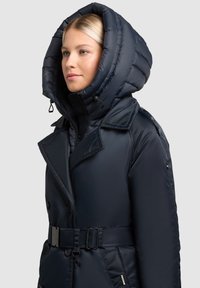 Navy Pufferjacke mit großer, gesteppter Kapuze, tailliertem Gürtel und doppelter Brustknopfleiste; hat eine glatte, glänzende Textur.