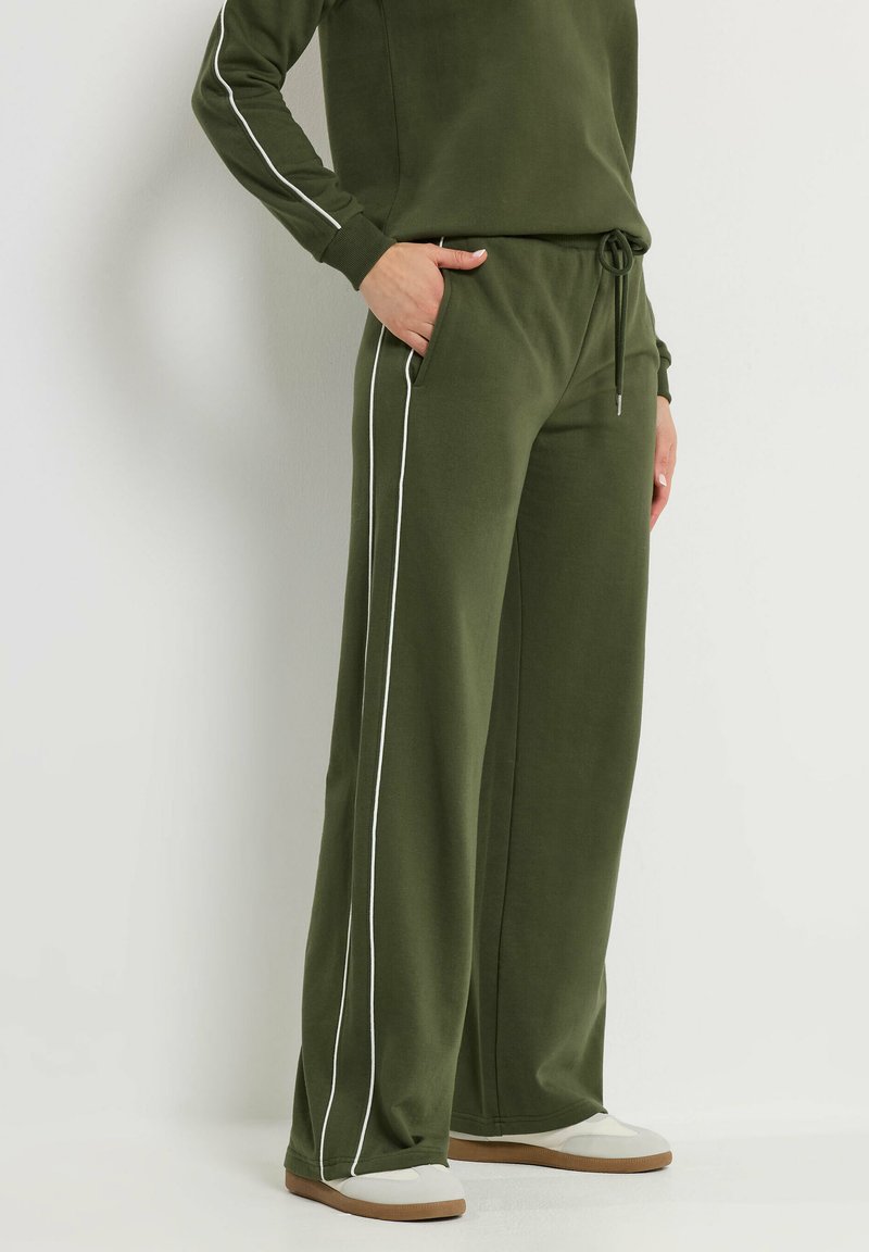 Person trägt olivgrüne, weit geschnittene Sweatpants mit weißen Seitenstreifen und passendem Sweatshirt, steht mit einer Hand in der Tasche.