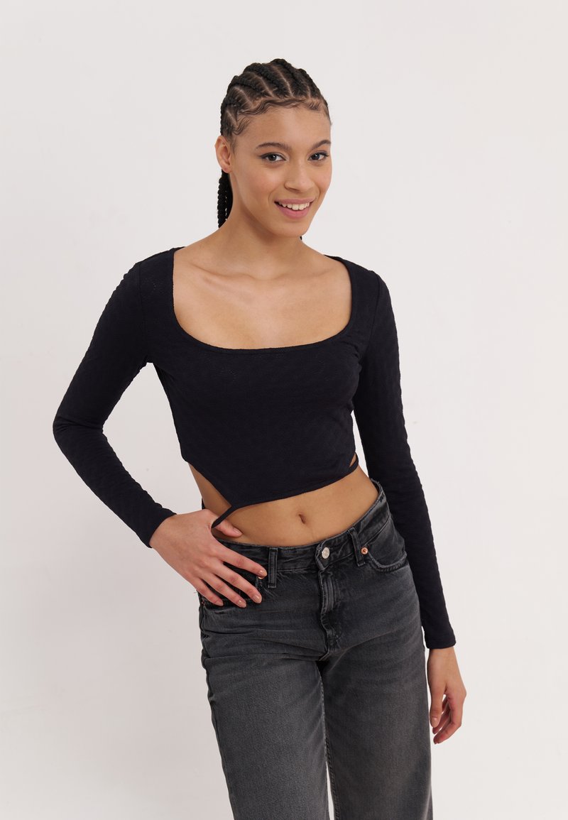 Black long-sleeve top with a square neckline and a cropped design, featuring textured fabric. Paired with high-waisted dark jeans.