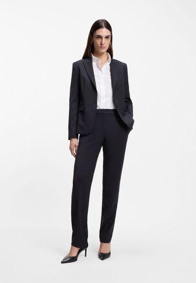 Femme portant un costume noir sur mesure avec un chemisier blanc et des chaussures à talons hauts noires, debout avec une main dans la poche sur un fond blanc.