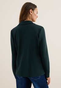 Donkergroene blazer met lange mouwen en een op maat gemaakte kraag, van gladde stof en een relaxte pasvorm, gecombineerd met blauwe denim jeans.