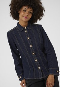 Veste en denim bleu foncé avec des boutons dorés, présentant un col, des détails de couture verticaux et des manches longues avec des poignets à boutons.