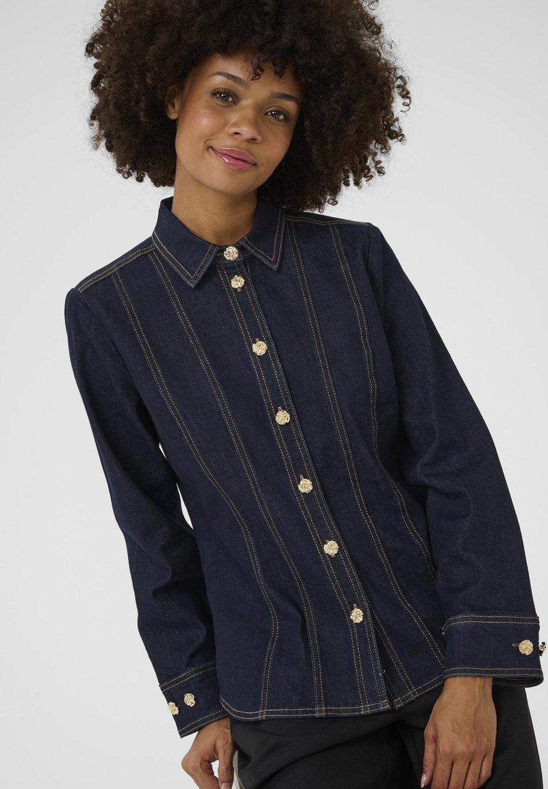 Námořnická denimová bunda se zlatými knoflíkovými zapínáními, s vertikálními švy a dlouhými rukávy. Má špičatý límec a strukturovaný střih.