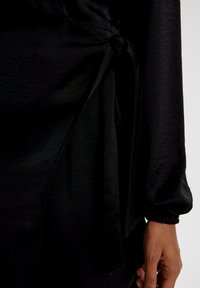 Schwarzes Wickelkleid mit strukturierter Oberfläche, das eine gebundene Taille und lange Ärmel mit elastischen Bündchen aufweist. Glattes, weiches Material.