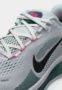 Ανοιχτό γκρι αθλητικό παπούτσι με μαύρο λογότυπο Nike. Διαθέτει ροζ και ασημί κορδόνια, επάνω μέρος από δίχτυ και υφή με σκούρα πράσινα στοιχεία.