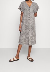 Šaty s květinovým potiskem mají výstřih do "V", krátké rukávy a zapínání na knoflíky vpředu. Látka je lehká; barevné schéma je černobílé.