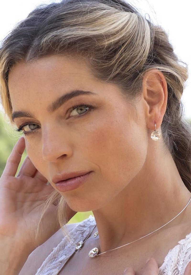 Zilveren sieraden omvatten ronde, doorzichtige oorbellen en een delicaat collier met kleine, afgeronde hangers. Het model heeft een lichte huid en neutrale make-up.