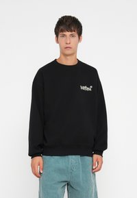 Zwarte crewneck sweatshirt met een klein wit "Wittopia" logo op de linkerkant van de borst. Zacht materiaal met een ontspannen pasvorm en geribbelde mouwen.