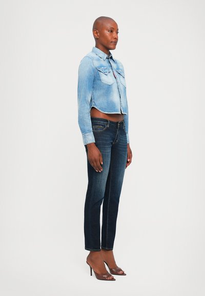Woman with shaved head wearing cropped light denim jacket, dark skinny jeans, and brown high-heeled open-toe shoes, standing sideways.