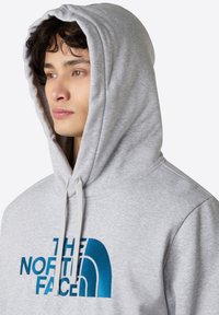 Grå hoodie i mjukt tyg, med en stor blå broderad logotyp. Inkluderar dragsko för huva och har en avslappnad passform.