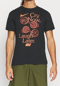 Camiseta de algodón negra con un diseño gráfico naranja que presenta pesas y el texto "Llora ahora, ríe después." Logo de Nike en la parte inferior. Mangas cortas.