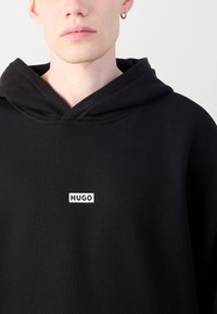 Schwarzer Kapuzenpullover mit einem weißen "HUGO"-Logo auf der Brust, weichem Material und standardmäßigem Kapuzen-Design.