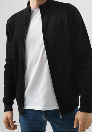 Mann trägt ein weißes T-Shirt, eine aufgezippt schwarze Jacke und blaue Jeans vor einfachem Hintergrund.