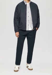Marine bomberjack over een wit-blauw gestreept shirt, gecombineerd met donkere broeken en witte sneakers met bruine accenten.