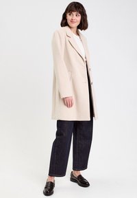 Manteau en mélange de laine beige avec revers crantés, deux poches avant et fermeture à trois boutons. Porté sur un haut blanc et associé à un jean foncé.