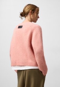 Różowy, puszysty sweter z okrągłym dekoltem, prążkowanym dołem i czarną metką z tyłu. Noszony z białą koszulą i zielonymi spodniami.