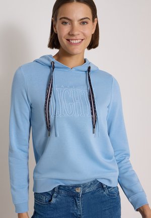 Femme souriante portant un sweat à capuche bleu clair avec des cordons à motifs et un jean bleu sur un fond clair uni.