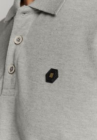 Grijze poloshirt van gestructureerde stof, met drie knopen en een zwart hexagonaal label met een horizontaal gestreept ontwerp.