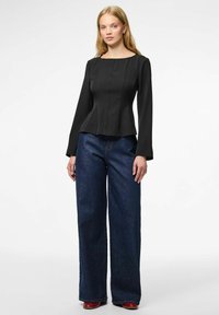 Maglietta nera a maniche lunghe con scollo aderente e orlo svasato, abbinata a jeans blu a gamba larga e scarpe rosse. Sfondo neutro.