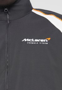 Μαύρο ελαφρύ μπουφάν με πορτοκαλί και λευκές λεπτομέρειες, με το λογότυπο της ομάδας McLaren Formula 1. Εμπρόσθιο φερμουάρ και σχέδιο με ψηλό γιακά.