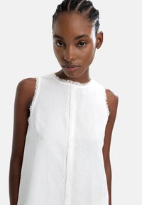 Biała lniana bluzka bez rękawów z postrzępionymi wykończeniami kołnierzyka i brzegów, z centralnym zapięciem na guziki i luźnym krojem.