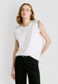 Valge ilma varrukateta topp, millel on studeeritud õlad ja franseerimise detailid, kombineeritud mustade pükstega. Sile kangas ja lõdva istuvusega.