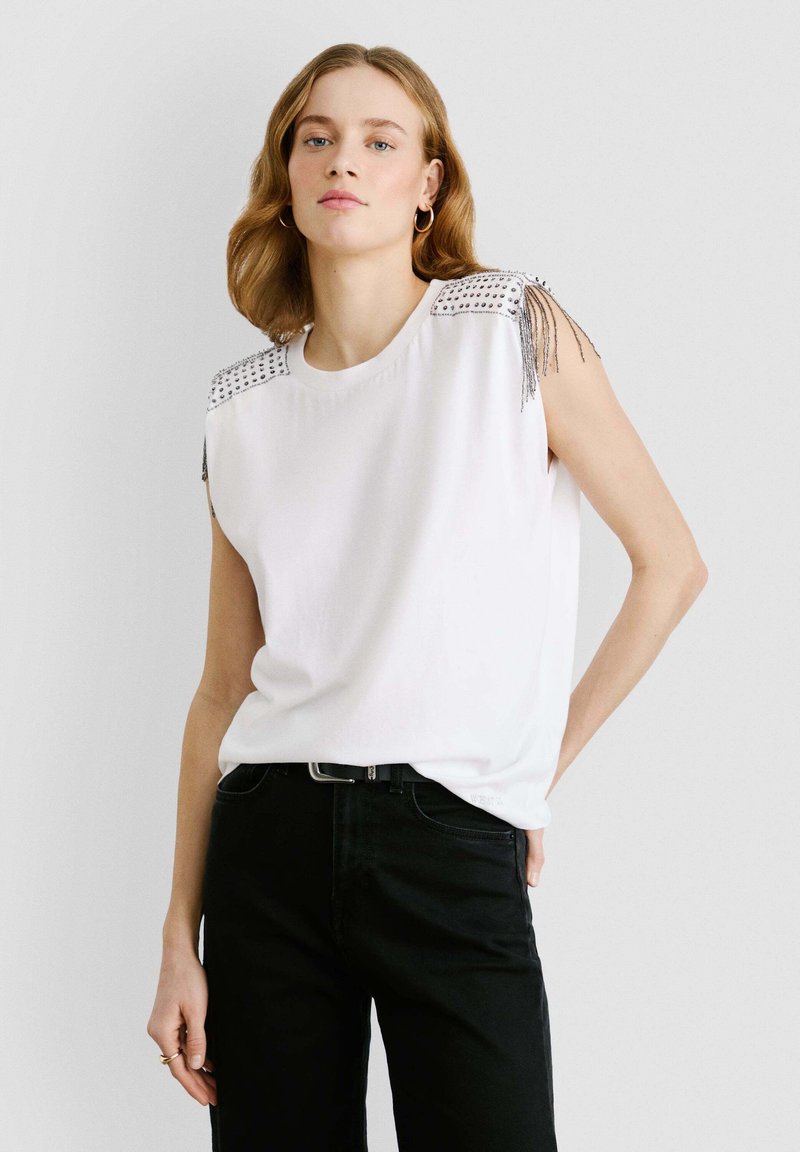 Valge ilma varrukateta topp, millel on studeeritud õlad ja franseerimise detailid, kombineeritud mustade pükstega. Sile kangas ja lõdva istuvusega.