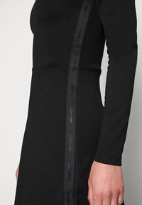 Czarna, długa sukienka z długim rękawem, o dopasowanym kroju, gładkiej fakturze i bocznym pasku z logotypem "Calvin Klein" w wytłaczanym napisie.
