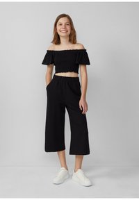 Schwarzes, schulterfreies Crop-Top mit rüschierten Ärmeln, kombiniert mit schwarzer, weit geschnittener cropped Hose; beide bestehen aus einem strukturierten Stoff. Weiße Sneakers.