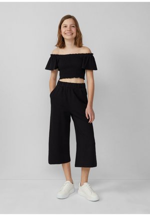 Zwarte off-shoulder crop top met ruches mouwen, gecombineerd met zwarte wijde cropped broek; beide hebben een textuurstof. Witte sneakers.