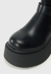 Botín negro de tobillo hecho de material sintético brillante, con punta redondeada, suela de plataforma con superficie texturizada y una correa decorativa.