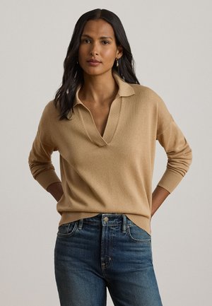 Femme aux longs cheveux foncés portant un pull camel à col en V avec col et un jean en denim bleu, debout les mains derrière le dos.