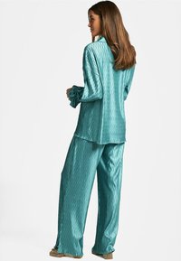 Conjunto de pijama de satén en color verde azulado, con textura estampada, mangas largas, corte holgado y pantalones de pierna ancha. Detalles de volantes notables en los puños.