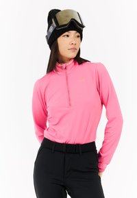 Dopasowany różowy top z długim rękawem i kołnierzem na pół zamek, wykonany z teksturowanej tkaniny, z złotym logo. Noszony z czarnymi spodniami i czarną czapką beanie z goglami.