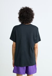 Pessoa com cabelo encaracolado de costas, vestindo uma t-shirt preta simples e calções roxos, contra um fundo claro e simples.