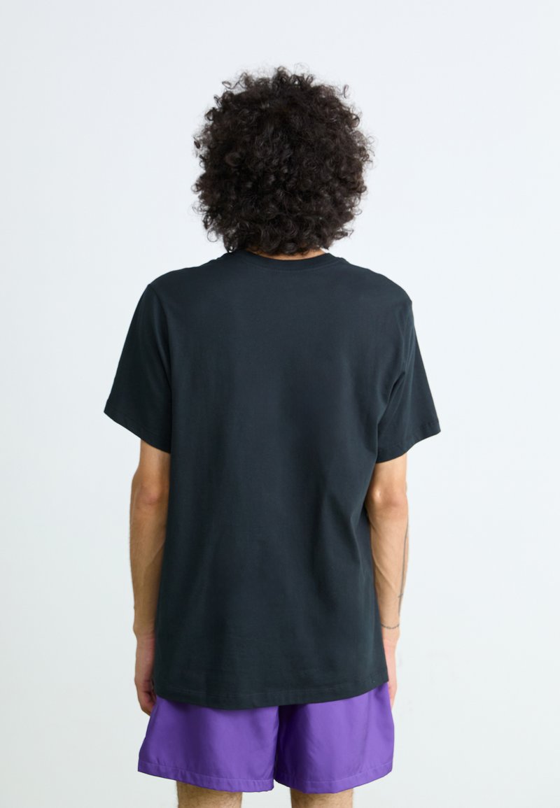 Persoon met krullend haar staat met de rug naar de camera, draagt een eenvoudige zwarte t-shirt en paarse shorts tegen een effen lichte achtergrond.