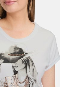 T-shirt blanc avec un graphisme monochrome d'une femme portant un chapeau de cowgirl, orné de perles et accentué par un design de collier scintillant.