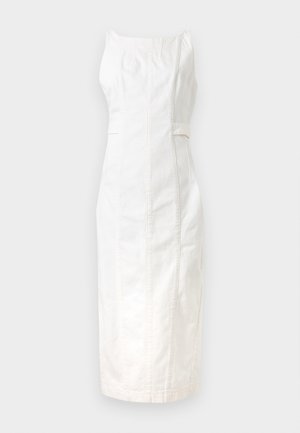 Žena s dlhými tmavými vlasmi, oblečená v bielych midi šatách bez rukávov a čiernych sandáloch s remienkami, stojaca pred jednofarebným svetlým pozadím.