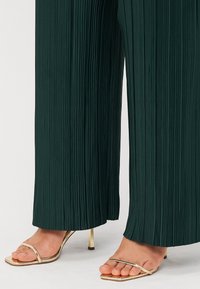 Pantalons larges plissés vert foncé assortis à des sandales à talons aiguilles dorées, montrant un détail de tenue élégant et stylé.