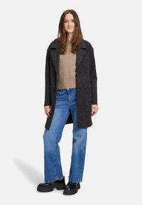 Donkergrijze lange jas met revers, gecombineerd met een lichtbruine sweater en blauwe wijde jeans. Glanzende zwarte platform schoenen maken de outfit compleet.