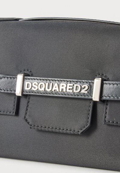 Gros plan sur un sac en tissu noir avec une sangle en cuir noir et des accessoires en argent portant le nom de la marque "DSQUARED2".