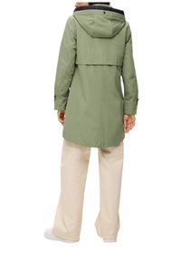 Imperméable vert olive avec une capuche noire, des manches longues et une coupe mi-longue. Le tissu semble lisse avec un léger éclat. Accents de boutons sur les manches.