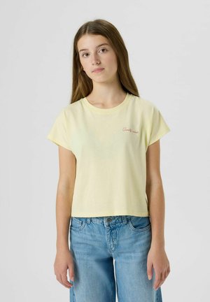 Junge Frau mit langen Haaren, die ein blassgelbes "Amalfi-Küste" T-Shirt und hellblaue Jeans trägt, vor einem einfarbigen hellen Hintergrund steht.