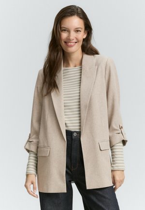 Femme souriante aux longs cheveux bruns portant un blazer beige sur une chemise rayée et un jean taille haute foncé, sur un fond uni.