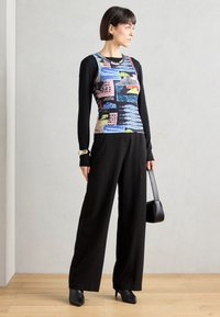 Colorful, patterned sleeveless top layered over a black long-sleeve shirt, paired with black wide-leg pants and pointed black heels.