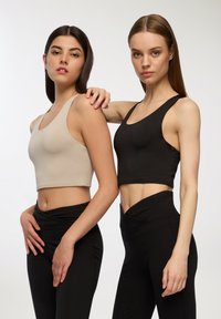 Due modelli indossano top corti aderenti; uno in beige e l'altro in nero. Entrambi i top hanno spalline larghe e una texture liscia. Sono abbinati a leggings neri a vita alta.