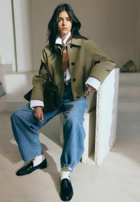 Žena v olivovej bunde, bielom tričku, hnedom šále, širokých modrých džínsoch, bielych ponožkách a čiernych mokasínach, sediaca na bielom kamennom bloku v interiéri.