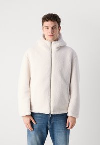 Bijela flis jakna s kapuljačom, prednjim zatvaračem i dugim rukavima. Mekana tekstura, opušteni fit, nosi se preko plavih traperica. Jednostavan, minimalistički dizajn.